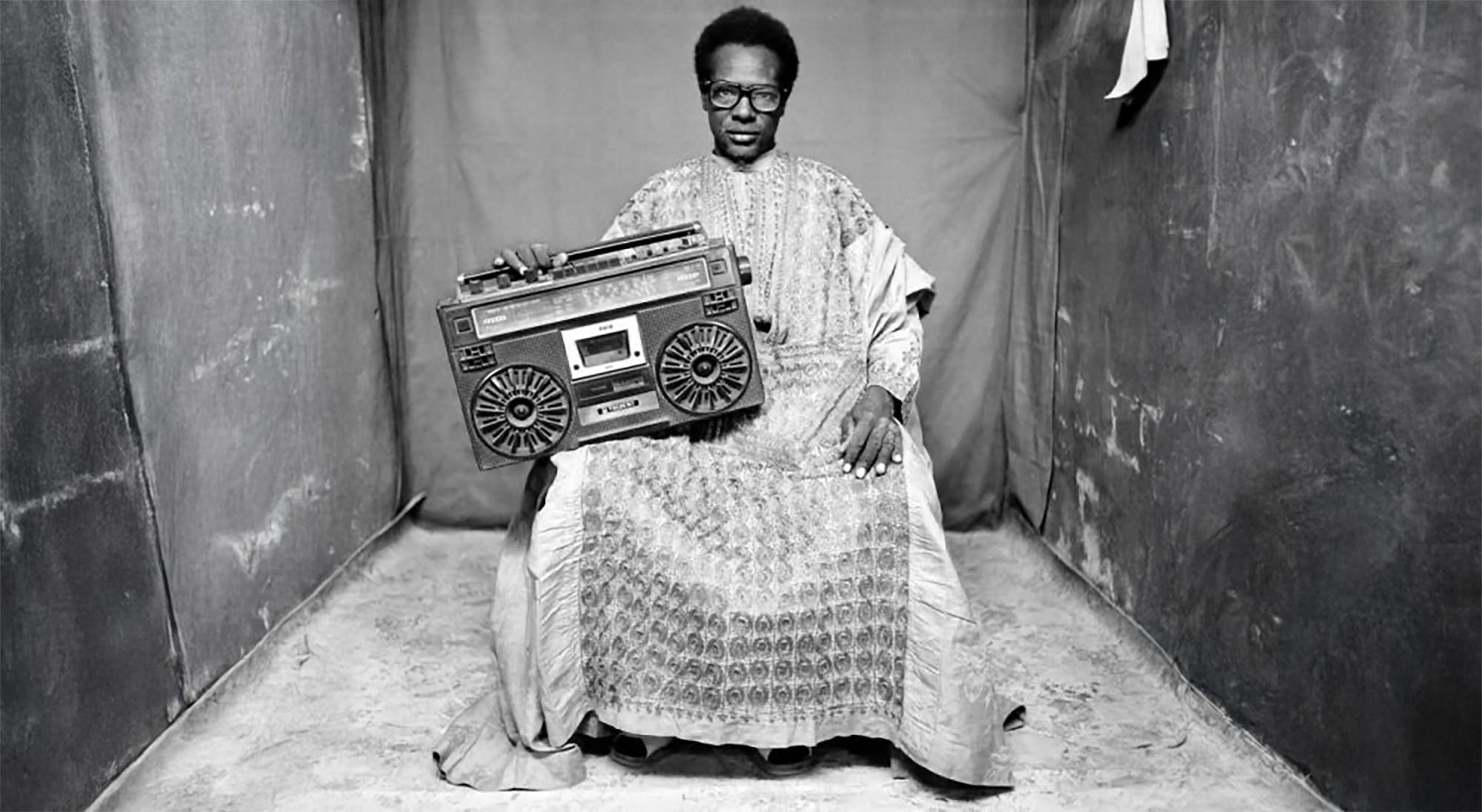 A black and white photograph of a seated man with glasses, holding a boombo